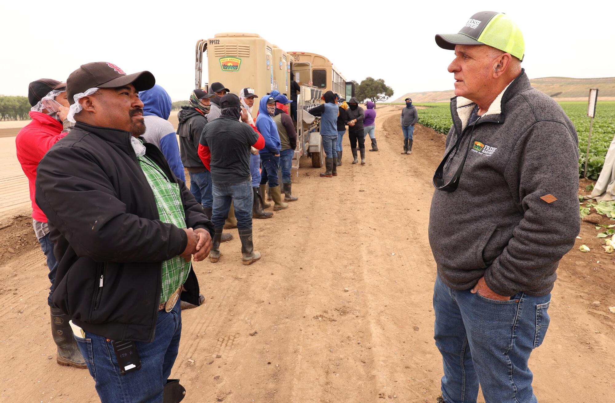 Two men talk in a field, while behind them farmworkers line up at hand-washing stations.