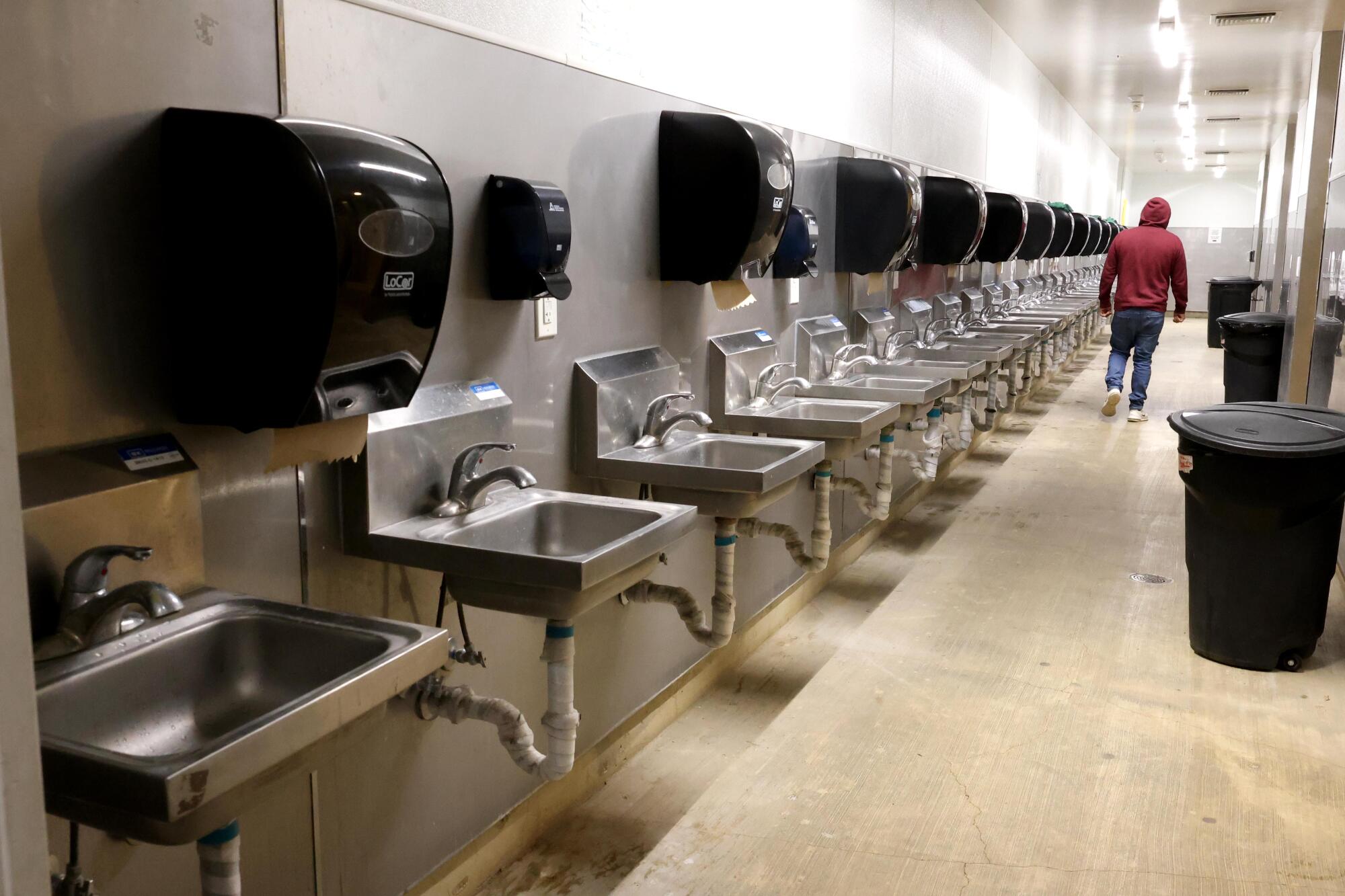 A man walks through a dormitory-style bathroom lined with stainless steel sinks.