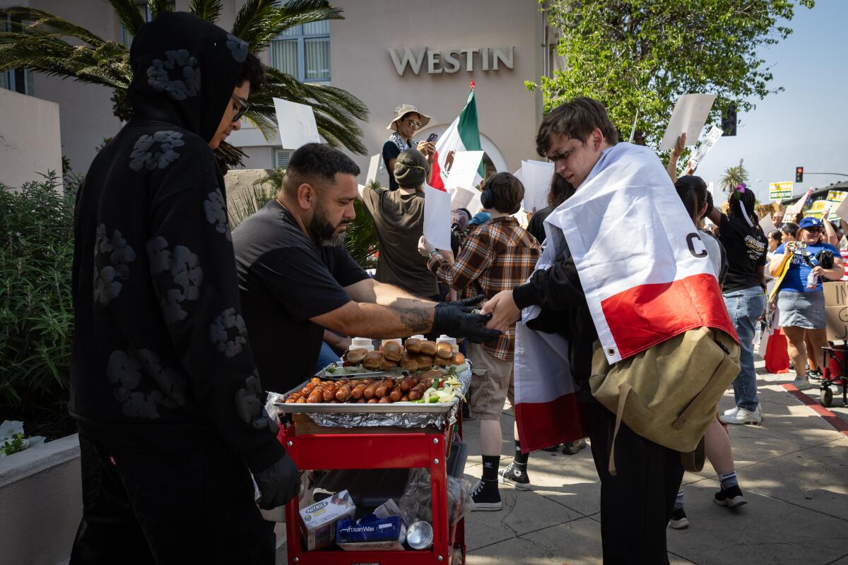A food vendor sells to a man with a Mexican flag draped over his shoulders as protesters stand in the background