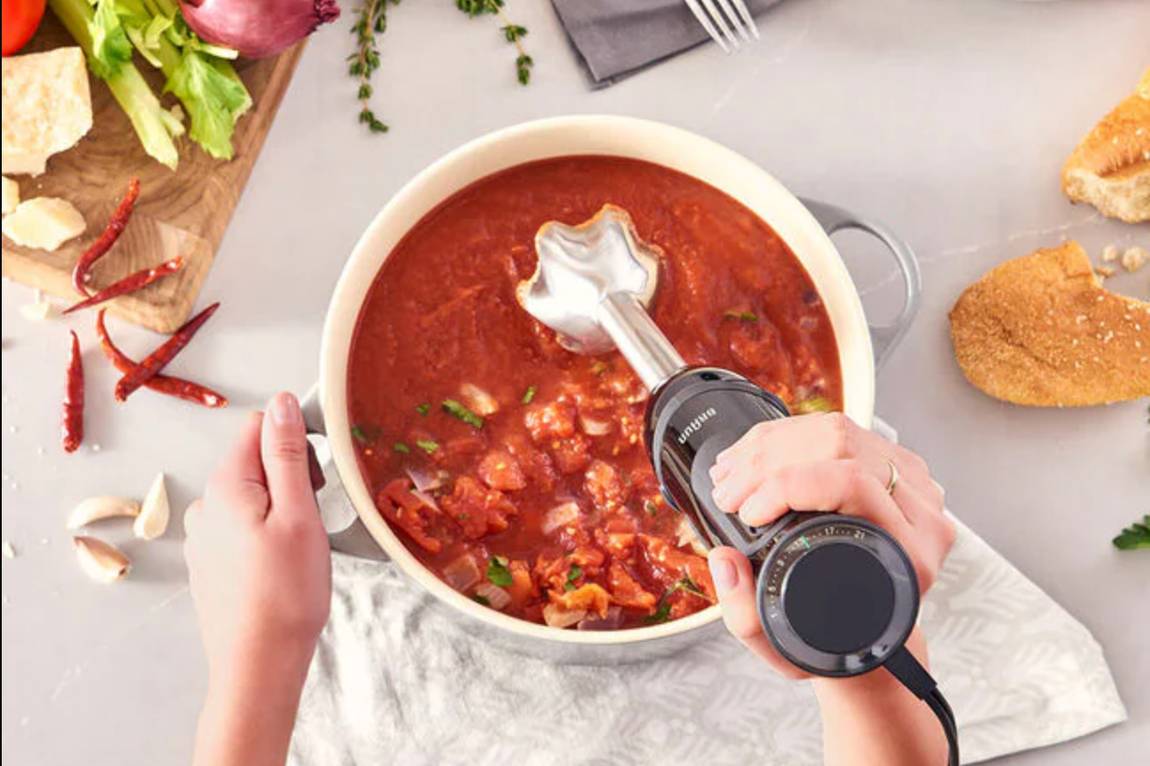 Overhead shot of hands holding a hand blender and stirring a bowl of a tomato-y broth.