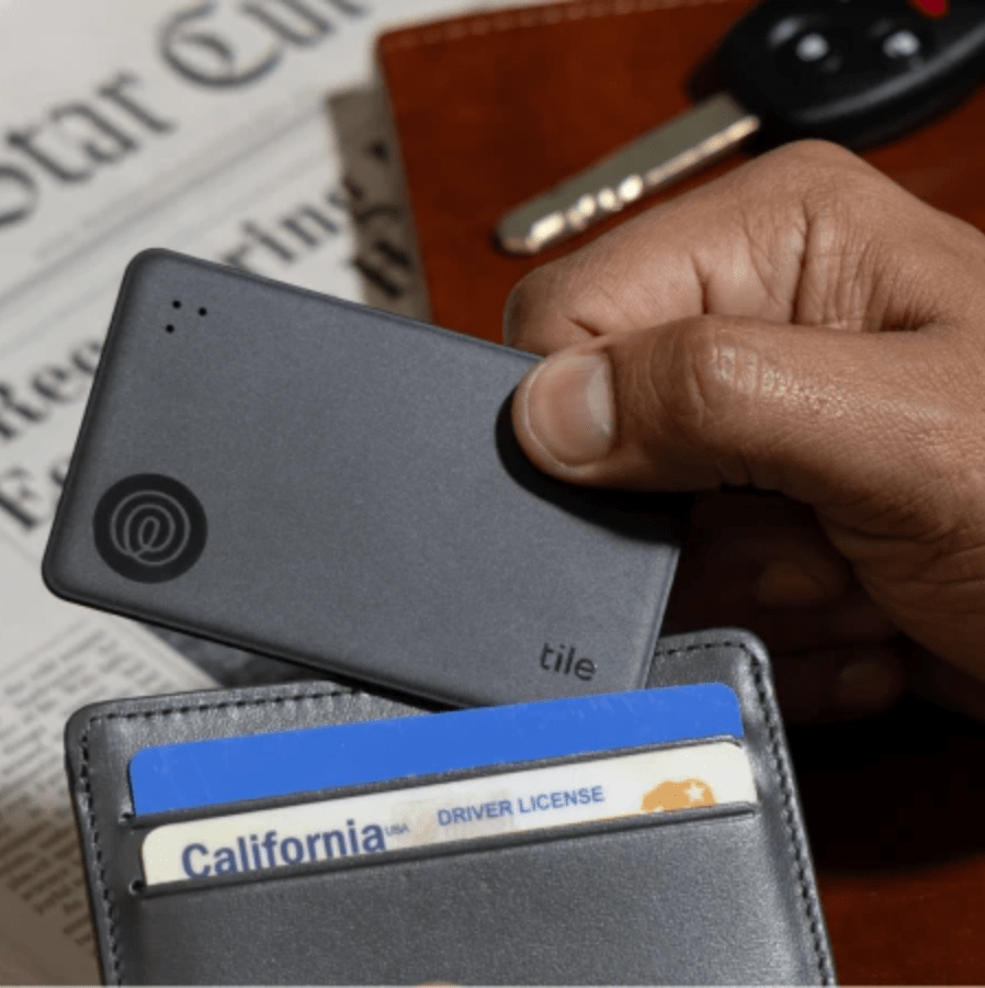 A hand placing a Tile Slim into a leather wallet with a newspaper in the background.