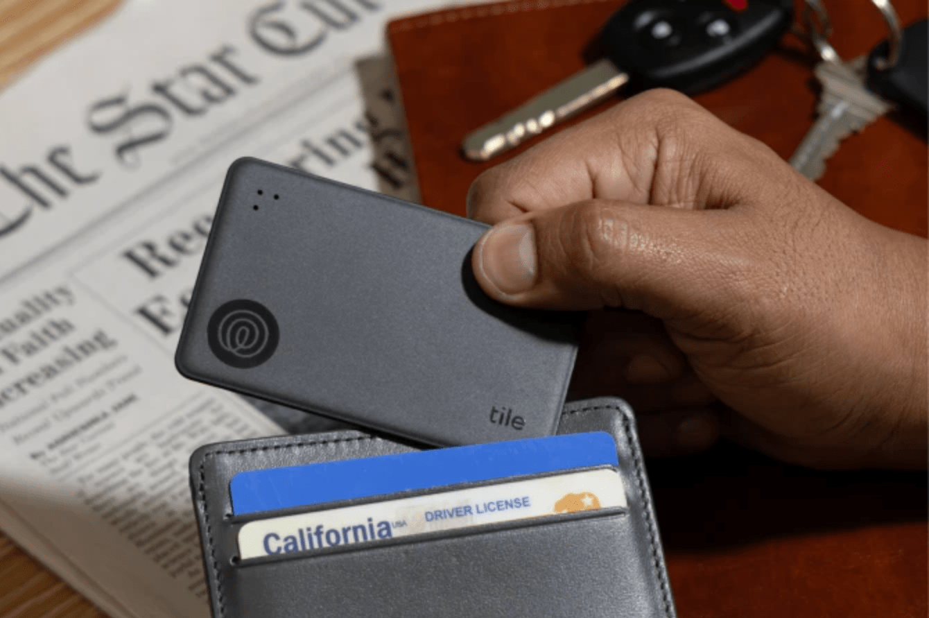 A hand placing a Tile Slim into a leather wallet with a newspaper in the background.