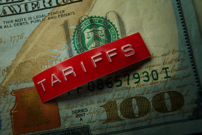 A red metal badge stamped with the word, tariffs, which is set atop a crisp one-hundred dollar bill.