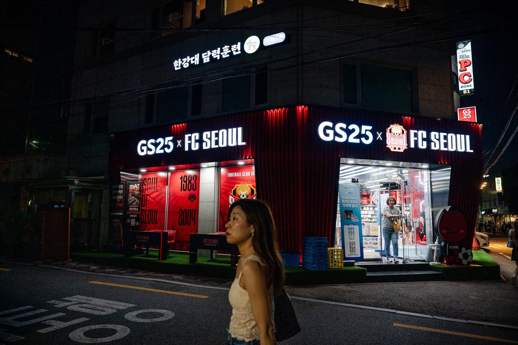 A woman walks by a building with a sign that reads GS25 x FC Seoul at night.